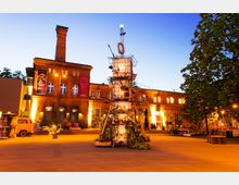 Roter Backsteinbau mit großem Schornstein, beleuchtet von warmem Licht, an einem offenen Platz während der Dämmerung. Im Vordergrund steht eine kunstvoll gestaltete, von Pflanzen umgebene Installation, umgeben von Bäumen und Sitzmöglichkeiten.