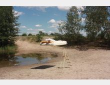 Ein sandiges Gelände mit einem kleinen Wasserbecken, umgeben von Bäumen. Im Vordergrund steht eine kleine gelbe Leiter, während schwebende Bettwäsche und Kissen in der Mitte des Bildes zu sehen sind.