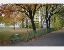 Ein kleiner Park mit herbstlichen Bäumen, einer grünen Parkbank und einem Metallzaun, im Hintergrund mehrstöckige Wohngebäude mit gelben Balkonen. Der Boden ist mit gefallenen Blättern bedeckt, und ein asphaltierter Gehweg führt durch den Park.