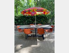 Ein Tisch mit einer grauen Steinplatte und sechs orangefarbenen Kunststoffstühlen steht auf einem gepflasterten Platz im Freien. Über dem Tisch befindet sich ein bunter Sonnenschirm mit Fanta-Werbung, im Hintergrund sind grüne Büsche und Bäume zu sehen.