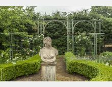 Steinbüste einer Frau auf einem Sockel in einem gepflegten Garten, umgeben von Buchsbaumhecken und einer grünen Metallpergola mit rankenden Rosen. Im Hintergrund dichter Bewuchs mit Bäumen und Sträuchern.