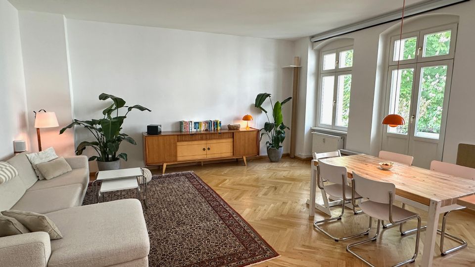 Helles Wohn- und Esszimmer mit Fischgrätparkett, großer beigefarbener Eckcouch auf gemustertem Teppich und Holzboden. An der Wand steht ein niedriges Sideboard mit Büchern und Lampe, daneben Zimmerpflanzen; rechts ein Esstisch mit Stühlen vor hohen Fenstern mit Blick ins Grüne.