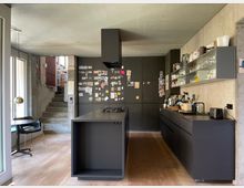 Moderne Küche mit dunklen Einbauschränken, einer zentralen Kochinsel und offenen Regalen an einer Betonwand. Im Hintergrund führt eine Betontreppe zu einem oberen Bereich, und links befindet sich eine Glasfront mit Blick nach draußen.