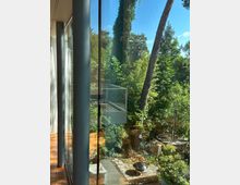 Blick durch eine bodentiefe Glasfront eines modernen Hauses auf eine Terrasse mit Glasgeländer und Holzdeck, umgeben von dichtem Grün. Im Garten sind hohe Bäume, Sträucher, Steinplatten sowie eine kleine Feuerstelle und ein rundes Becken mit Metallrohr zu sehen.