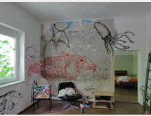 Ein renovierungsbedürftiger Raum mit Wandmalereien von Meerestieren, darunter ein großer roter Fisch, ein Mantarochen und ein Tintenfisch, auf einer abgenutzten Wand. Vor der Wand stehen zwei Stühle und ein kleiner Tisch; durch eine offene Tür ist ein Schlafzimmer mit einem orangefarbenen Bettlaken sichtbar.