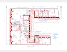 Grundriss eines Gebäudes mit roter Markierung der Wände, technischen Anmerkungen und Symbolen für Türen, Fenster und Sanitäranlagen. Die Zeichnung zeigt mehrere Räume, ein zentrales Treppenhaus und Beschriftungen für Installationen und bauliche Details.