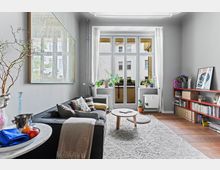 Wohnzimmer mit großen Fenstern und einem Blick auf den Innenhof. Der Raum ist mit einem grauen Sofa, einem runden Holztisch, einem hellen Teppich und einer Bücherwand eingerichtet; auf den Fensterbänken stehen mehrere Zimmerpflanzen.