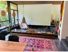 Modernes Wohnzimmer mit großen bodentiefen Fenstern, die den Außenbereich mit viel Grün zeigen. Die Einrichtung umfasst einen hängenden Korbsessel, ein tiefes Sofa mit Holztisch, einen bunten Teppich mit geometrischem Muster und Pflanzen in Töpfen; die Decke ist mit Holzverstrebungen verkleidet.