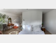 Helles Schlafzimmer mit weiß gestrichenen Wänden, einem Doppelbett mit weißen Kissen und Bettwäsche. Im Raum befindet sich ein kleiner Holzschreibtisch mit Spiegel, ein orangefarbener Sitzsack, ein grauer Sessel, eine Stehlampe sowie ein Teppich mit orientalischem Muster auf hellem Holzboden.