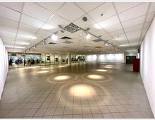 Leerer Innenraum in einem Einkaufszentrum mit geflieste Böden, weißer Deckenverkleidung mit integrierten Leuchten und einer Glasfront auf der linken Seite, hinter der Geschäfte und Kunden sichtbar sind. Im Hintergrund befinden sich Ausgänge und Türen.