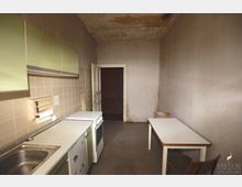 Schmale, veraltete Küche mit abgenutzten Wänden und Decke, beige Fliesenrückwand und grünen Hängeschränken. Auf der rechten Seite stehen ein Tisch und zwei Holzstühle, auf der linken eine einfache Küchenzeile mit Spüle, Herd und Kühlschrank.