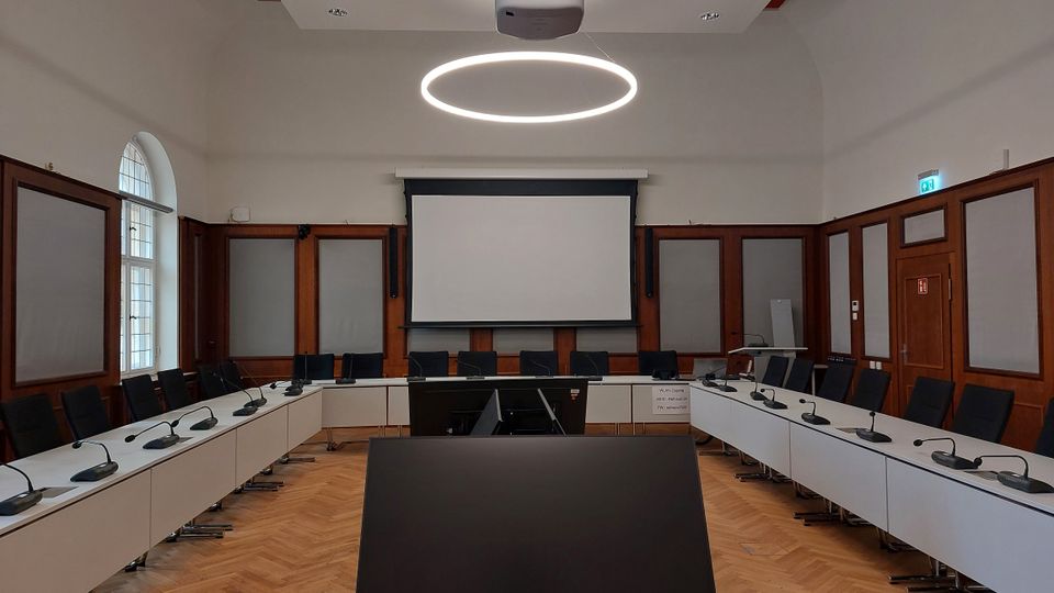 Konferenzraum mit oval angeordneten Tischen, Mikrofonen auf den Arbeitsplätzen und schwarzen Stühlen. Im Hintergrund befindet sich eine große Leinwand, umgeben von holzgerahmten Akustikpaneelen; die Decke ist hoch mit einem modernen, kreisförmigen Leuchtring als Beleuchtung.
