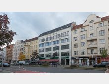 Straßenszene mit mehrstöckigen Wohn- und Geschäftsgebäuden im typischen Berliner Altbaustil, mit einer Fassade mit großen Fenstern und Verzierungen auf einem Gebäude. Im Erdgeschoss sind Geschäfte wie "Woolworth" erkennbar, davor geparkte Autos, Bäume und eine Straße mit Fahrradweg.