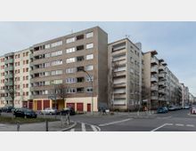 Wohnblock in einer Stadtansicht, bestehend aus mehreren mehrstöckigen Gebäuden mit schlichten Fassaden und Balkonen. Im Vordergrund befindet sich eine Straßenecke mit geparkten Autos, Gehwegen und einigen kahlen Bäumen.