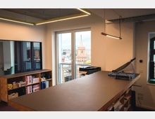 Moderner Büroraum mit Holzschreibtisch, Regalen und Büroausstattung. Durch große Fenster ist ein Blick auf eine historische Backsteinarchitektur zu sehen, einschließlich eines Kirchturms.