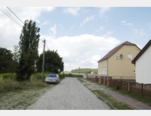 Eine ruhige Dorfstraße mit Kopfsteinpflaster, gesäumt von einer Wiese auf der linken Seite und Wohnhäusern mit Zäunen auf der rechten Seite. Ein geparktes Auto steht am Straßenrand, im Hintergrund sind Bäume und Felder erkennbar.