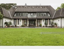 Großes, traditionelles Wohnhaus mit Satteldach, weißer Fassade und Steinpfeilern, umgeben von einem gepflegten Garten mit Rasenfläche. Vor der Terrasse stehen mehrere Topfpflanzen, Gartenmöbel und Sonnenschirme.