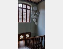 Holztreppe in einem Altbau mit großen Fenstern, darunter ein mehrteiliges buntes Glasfenster in Blumenform. Eine moderne Hängelampe mit kugelförmigen Leuchtelementen hängt von der dekorativ verzierten Decke.