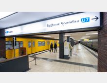 U-Bahnhof mit gelbem Zug der Linie U7, der gerade einfährt. Auf dem Bahnsteig sind Passagiere sowie ein Fahrkartenautomat und Wegweiser zu sehen, die umliegenden Wände sind mit dunklen Fliesen gestaltet.