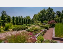 Ein gepflegter Garten mit gepflasterten Wegen, umgeben von dichten grünen Hecken, bunten Blumenbeeten und Ziergräsern. Im Hintergrund steht eine Holzbank zwischen Bäumen und Sträuchern, die eine ruhige Atmosphäre schaffen.