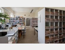 Großes Büro mit Regalen voller Aktenordner und gestapelten Papierdokumenten. Zwei Schreibtische mit Computern, ein Drucker und Pflanzen stehen im Vordergrund, während eine Tür zu einem weiteren Raum im Hintergrund sichtbar ist.