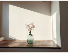 Ein schlichtes Arrangement mit einer grünen Glasvase, die getrocknete Zweige enthält, steht auf einer Holzoberfläche vor einer cremeweißen Wand. Sonnenlicht fällt durch ein Fenster und erzeugt geometrische Schatten auf der Wand und der Oberfläche.