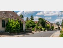 Straßenszene in einem Vorort mit Einfamilienhäusern; die Häuser haben Ziegeldächer, helle Fassaden und sind von Gärten mit dichter Vegetation umgeben. Eine asphaltierte Straße mit gepflastertem Gehweg und Straßenlaternen führt entlang der Häuser.