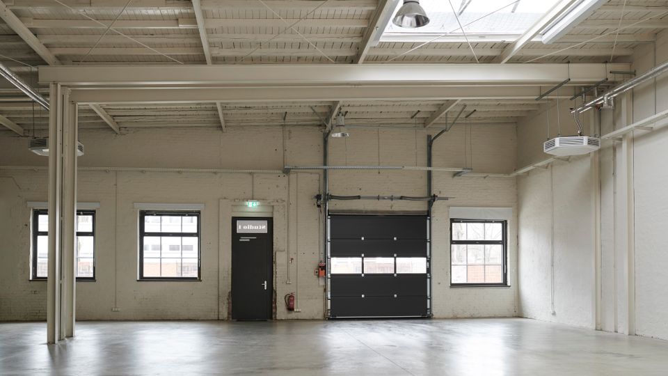 Empty industrial warehouse interior with white-painted brick walls, a polished concrete floor, and exposed beams, ducts, and hanging lights. Several rectangular windows line the wall beside a door and a large black roll-up garage door under a skylight.