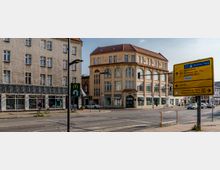 Straßenszene mit einem beige-gelben, mehrstöckigen Gebäude im Eckhaus-Stil mit großen Fenstern und historischer Fassade, daneben ein Wohn- und Geschäftsgebäude. Im Vordergrund ein gelbes Straßenschild mit Entfernungsangaben und eine McDonald's-Beschilderung.