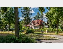 Ein Wohnhaus mit rotem Satteldach und heller Fassade, umgeben von Bäumen und einer gepflegten Hecke. Im Vordergrund führt eine gepflasterte Straße mit Straßenschildern durch eine grüne, parkähnliche Umgebung, ein Radfahrer fährt auf dem Weg.