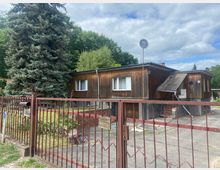 Einfamilienhaus in Holzbauweise mit Flachdach, umgeben von Garten und einer Einfahrt aus Betonplatten. Im Vordergrund ein roten Metallzaun mit Briefkasten; rechts ein Nebeneingang mit kleiner überdachter Treppe. Im Hintergrund Bäume und blauer Himmel mit Wolken.
