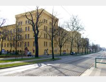 Gelbes, mehrstöckiges historisches Gebäude mit rechteckigen Fenstern und Zinnen am Dachrand, flankiert von kahlen Bäumen. Davor verläuft eine Straße mit Straßenbahnschienen und Gehwegen.