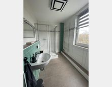 Modernes Badezimmer mit begehbarer Dusche mit Glastüren, Waschbecken an einer türkis gefliesten Wand, wandhängendem Heizkörper und grauen Bodenfliesen. Ein großes Fenster mit Jalousien sorgt für Tageslicht.