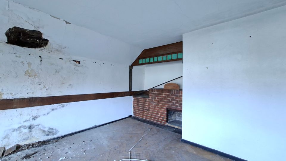 Innenraum eines leerstehenden, stark beschädigten Zimmers mit abblätterndem Putz, Rissen in der Decke und einem großen Loch in der linken Wand. Rechts steht eine gemauerte Ziegelkonstruktion mit offenem Kamin oder Nische, darüber ein Holzaufsatz mit schmalen grünen Glasfenstern.