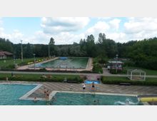 Ein Freibad mit mehreren Becken, darunter ein großes rechteckiges Schwimmbecken und ein flacherer Bereich im Vordergrund, umgeben von Grünflächen und Bäumen. Am Beckenrand stehen gelbe Bänke und eine kleine, pink-weiße Hütte, während sich im Hintergrund Wälder erstrecken.