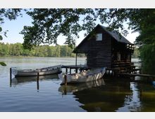 Holzhütte am Ufer eines Sees mit einem kleinen Steg, an dem zwei Ruderboote befestigt sind; im Hintergrund eine von Bäumen gesäumte Landschaft.