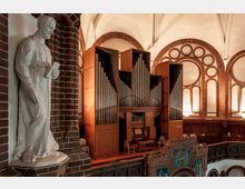 Innenansicht einer historischen Kirche mit großen Orgelpfeifen in einem hölzernen Gehäuse. Links ist eine weiße Steinstatue vor einer roten Backsteinwand zu sehen, während die Umgebung durch Bögen und verziertes Mauerwerk geprägt ist.