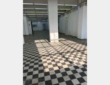 Ein leerer Innenraum mit rechteckigem Grundriss, schwarz-weiß kariertem Fliesenboden und weißen Wänden. Die Decke ist mit langen, weißen Leuchtstofflampen versehen, und vereinzelt sind Türen und Wandnischen sichtbar.