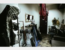 Small, cluttered backstage dressing room with white textured walls, wooden floorboards, and a vanity shelf beneath a wall mirror. Costumes and props hang from hooks and a ceiling rack, with instrument cases and frames stacked along the walls near a black door and ornate mirrors.