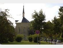 Eine historische Kirche mit achteckigem Grundriss und gotischen Spitzbogenfenstern, umgeben von Bäumen und Grünflächen. Im Vordergrund sind ein Verkehrszeichen und eine Baumreihe entlang einer Straße sichtbar.