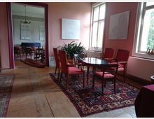 Ein historisch eingerichtetes Zimmer mit Holzboden, einem runden Holztisch und vier roten Polsterstühlen, die auf einem orientalischen Teppich stehen. Große Fenster lassen Tageslicht herein, während Kunstwerke an den rosafarbenen Wänden hängen; im Hintergrund ist ein angrenzender Raum mit weiterer Möblierung und einem Kronleuchter sichtbar.