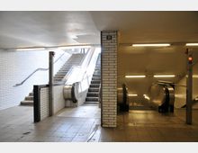 Unterirdischer Zugang mit gefliesten weißen Wänden, einer Treppe in der Mitte und Rolltreppen auf beiden Seiten. Der Bereich ist beleuchtet durch Deckenlampen, und der Boden ist mit hellen Fliesen ausgelegt.