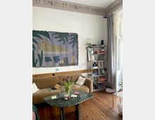 Wohnzimmer mit Holzboden, einem beigen Sofa und einem grünen Marmortisch, auf dem persönliche Gegenstände stehen. An der Wand hängt ein dekorativer Wandteppich mit abstrakten Mustern, daneben ein Regal mit Büchern und Pflanzen. Rechts im Bild befindet sich ein Fenster mit hellen Vorhängen.