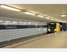 U-Bahn-Station "Frankfurter Tor" mit hellblauen Wandfliesen und einem dunkelblauen Streifen mit Stationsnamen. Rechts fährt ein gelber U-Bahn-Zug in die Station ein.