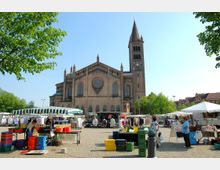 Ein belebter Marktplatz mit Verkaufsständen und bunten Kisten vor einer großen Kirche im neoromanischen Stil mit Spitzbogenfenstern, einem Rosettenfenster und einem hohen Turm mit Uhr. Im Hintergrund sind Bäume und Ziegelhäuser erkennbar.