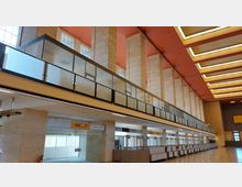 Großer, leerer Flughafen-Terminalbereich mit hohen Säulen aus hellem Stein, einer roten Decke und langen Reihen von Schaltern. Im oberen Bereich befindet sich eine verglaste Galerie mit undurchsichtigen Fenstern.