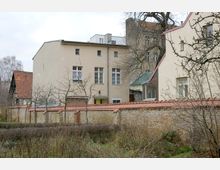 Hinterhof mit mehreren Wohngebäuden in heller Putzfassade und Ziegeldächern, umgeben von einer niedrigen Mauer aus Backstein mit roten Dachziegeln. Im Vordergrund sind kahle Bäume und ein verwilderter Garten zu sehen.