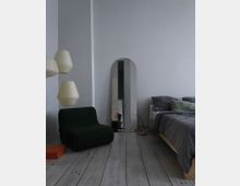 Minimalistisch eingerichtetes Schlafzimmer mit hellem Holzboden, einem dunkelgrünen Sessel, einem bodenlangen, abgerundeten Spiegel, einer Hängelampe mit Papieroptik und einem Bett mit grauer Bettwäsche. Auf dem Boden liegen ein oranger Karton und ein aufgeschlagenes Buch.