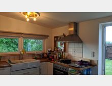 Helle, gemütliche Küche mit Holz-Arbeitsplatten, Doppelspüle, einem Fenster mit Blick auf grüne Bäume und einer offenen Tür zum Garten. Eine Dunstabzugshaube aus Edelstahl befindet sich über einem Herd, daneben Regale mit Gewürzen und Küchenutensilien.