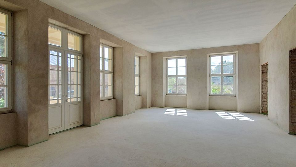 Großer, leerer Innenraum im Rohbau mit verputzten Wänden, Betonboden und hoher Decke. Mehrere hohe Sprossenfenster sowie eine verglaste Doppeltür lassen viel Tageslicht herein; rechts ist ein offener Durchgang mit sichtbarem Mauerwerk.